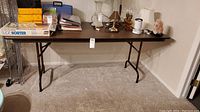 Wide shot of the 6' folding table against a beige wall, showing its entirety including folding metal legs, carpet floor, and various items on top.