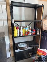 Photo of first black metal garage shelving unit showing 4 wide shelves with various cleaning and maintenance bottles on middle shelves, clock and other items on lower shelf. Shelving dimensions approximately 36x15.5x72 inches.