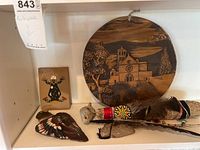 Overview photo showing all items on white shelf including large round carved wood artwork, small painted artwork, wooden bird sculpture, and decorated horn pieces.