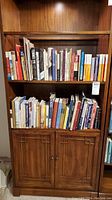 Full view of the wooden bookshelf containing two shelves filled with books and two decorated cabinet doors below. Close to floor, beige tiled flooring underneath.