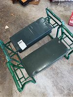 Two green metal framed garden kneelers with padded vinyl kneeling surfaces placed side by side on a concrete floor.