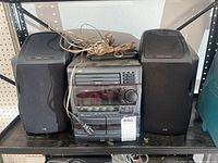 Photo of JVC stereo system including main 3 disc CD and cassette player unit flanked by two matching speakers on metal shelving.