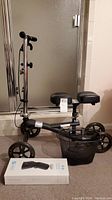 Full side view of black knee scooter with adjustable handlebar, two padded knee rests, and front mesh basket. Knee brace box placed in front on floor.