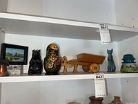Photo showing the entire row of assorted knick knacks on a white shelf including a wooden wagon, nesting doll, black bear statue, teal cat, green glass insulator, miniature framed painting, and copper bell.