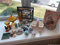 Full view of knick knacks and paper weights arranged on a window ledge including glassware, small ceramics, paper weights, framed mirror, and decorative stones.