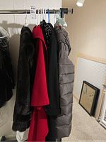 Five ladies coats and jackets hanging on a clothing rack, colors include black, red, and grey. Various styles: faux fur, wool, puffer, and satin visible.