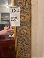 Close-up photo of the mirror's ornate frame showing carved scroll and leaf patterns with gold-toned highlights.