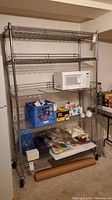 Full view of the metal wire shelving unit holding miscellaneous items including a microwave, crate, and various household goods. The shelving unit has five shelves and caster wheels at the base.
