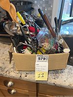 Box containing assorted kitchen utensils and gadgets including wooden spoons, handheld tools, and a glass jar with small items.