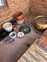 Overall view of different sized ceramic plant pots arranged on a porch corner with brick wall background, including large and small pots in various colors and shapes.