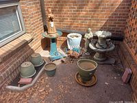 Overview photo showing most of the pots, statues, and outdoor decorative items arranged on the porch corner with brick walls. Visible items include various ceramic pots, a dog statue, a meditating statue, and concrete fountains with scattered dry leaves and pine needles.