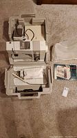 Open two-piece beige case showing top compartment with power supply/foot pedal and bottom holding extension table and accessories; dust cover at right