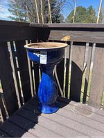 Tall blue ceramic planter on outdoor wooden deck corner, showing full height and shape with soil inside.