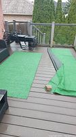Two pieces of green astro turf laid flat on an outdoor wooden deck with glass railings, one piece is partially rolled showing black backing, brick holding one corner down.
