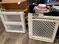 Two white baby gates, one with diamond mesh and one with circular hole panel, on floor with shelves and items in background.