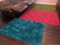 Three rectangular shag carpets on dark hardwood floor, colors teal blue, bright pink, and lime green. Part of a brown leather sectional sofa visible.