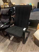 Side angle view of black upholstered chair with padded seat and wood frame, located indoors on wood flooring near bed and other chairs.