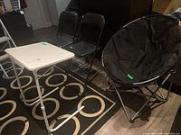 View of two black folding chairs, one round black folding chair, and one white plastic folding table arranged on a patterned rug.