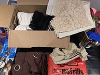 Box with folded beige lace curtains, black and white fabric curtains inside, brown curtain with grommets, and other assorted fabric curtains and table cloth.