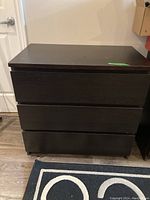 Front view of three-drawer dark wood dresser on a rug with circular pattern, showing flat drawer fronts and smooth wood finish.