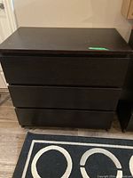 Front angle view of a 3 drawer dresser in dark wood finish on a wooden floor with black and white patterned rug. Shows the drawer fronts without handles.