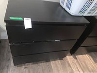 Front view of black wood dresser with three drawers and no visible handles, on a dark wood floor.