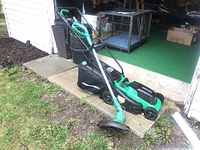 Electric lawn mower alongside grass trimmer positioned outdoors on a patio edge beside garden soil.