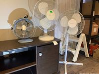 Photo of three electric fans identified by seller: small desk fan (13 inch), medium pedestal fan (24 inch), and large pedestal fan (42 inch) arranged on and beside a low cabinet in basement storage.