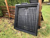 Black plastic square greenhouse base with gridwork, leaning against a wooden fence outdoors.