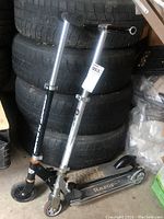 Two kids scooters leaning against stacked tires in storage shed, one Razor and one Street Runner brand, showing adjustable handlebars and wear