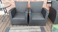 Two dark grey resin wicker-look outdoor armchairs on wood deck against brick wall next to round glass-top table