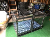 Large coated metal pet crate with plastic tray insert visible on the green floor in a shed environment, showing overall structure and doors.