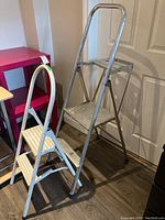 Two metal step ladders placed near a door and pink shelf, showing relative size and condition.