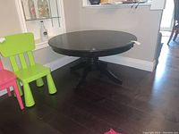 Table shown with round dark wood top, central pedestal and four legs, placed on dark wooden floor next to colorful chairs.