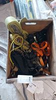 Box containing various tie straps and ropes including yellow ratchet straps, orange and black tie straps, with visible metal ratchet components.