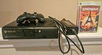 Black Xbox 360 console with wired controller on top, visible power supply and sealed EndWar game case in background.