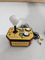 Photo showing Frogger branded plug and play TV video game unit, yellow background with frog graphic, joystick with white round top and two white buttons, RCA output cables visible.