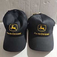 Front view of two identical John Deere black baseball caps showing the yellow embroidered logo and "JOHN DEERE" text.