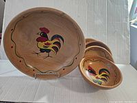 Full view of several wooden bowls stacked together, all featuring a painted colorful rooster design in the center and a decorative border around the rim.