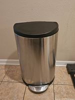 Front view of a stainless steel trash can with a black lid and foot pedal on tile floor.