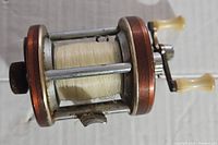 Side view of vintage Heddon Heritage Mark III fishing reel with fishing line on spool, showing metal frame between bronze-colored side plates and dual handle crank