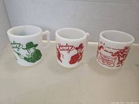 Photo of three translucent white milk glass cups with printed Western-themed graphics of Hopalong Cassidy in green and red and Davy Crockett in red.