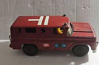 Side view of vintage red toy ambulance showing white medical cross on roof, black wheels, yellow driver figure and some surface wear.