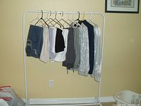 Full view of 8 pants and 3 sweaters hung on a white metal clothing rack against a wall, showing a variety of styles and colors.