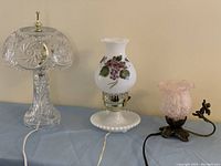 Three lamps displayed together: clear pressed glass lamp, white floral glass lamp, cranberry glass lamp with metal base and handle.