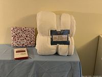 Photo showing folded white Kirkland king size plush blanket, floral patterned fitted sheet, and standard pillowcases in packaging.