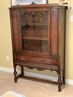 Full front view of the antique wooden china cabinet showing the glass door with decorative woodwork and turned legs.