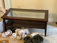 Wooden coffee table with dark wood, glass top insert, lower wooden shelf, and four legs, shown with surrounding items on carpeted floor.