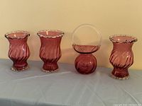 Four cranberry glass items: three vases and one basket displayed side by side on table covered with cloth