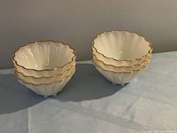 Six white glass dessert bowls with gold trim stacked in two stacks of three, showing scalloped, wavy edges and fluted sides.